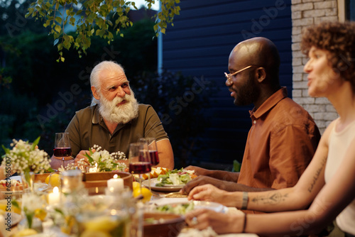 Bearded senior man looking at young African American guy during chat while sitting by table served with homemade food and having dinner