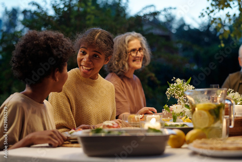 Cute African American girl looking at her brother during communication while both sitting by served table during outdoor family dinner