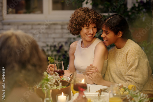 Happy girl sitting next to her smiling mother by dinner table served with homemade food and drinks during chat while enjoying summer evening