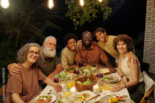 Cheerful intercultural family of three generations sitting by served table during outdoor dinner in the evening by their country house