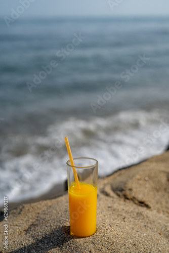 a glass of orange juice with a straw stands on the sand against the backdrop of the sea