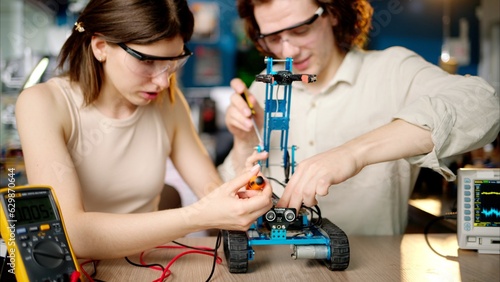 Two young happy engineers fixing a mechanical robot car in the workshop, using VR virtual reality headsets