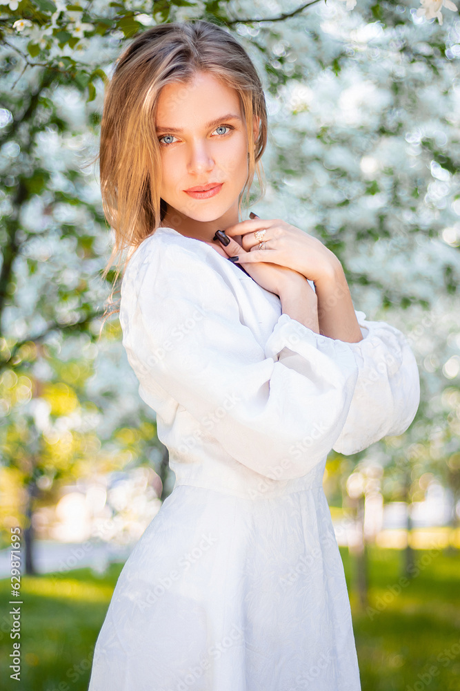 Portrait of a young attractive woman in a white dress against the backdrop of blooming apple trees. The girl poses against the background of flowers in the spring park. Portrait of a young attractive woman in a white dress against the backdrop of blooming apple trees. The girl poses against the background of flowers in the spring park.
