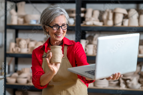 Woman pottery artist using laptop in art studio showing her earthenware products, selling online. photo