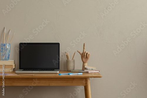 Aesthetic composition of simple office interior with copy space, wooden desk, computer, books, sculpture, glass cup with pencils, brown wall and personal accessories. Home decor. Template.