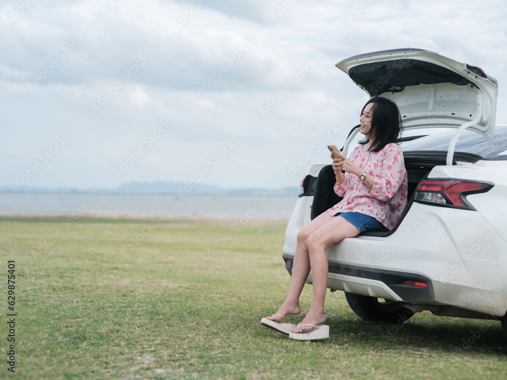 Asian woman using a smartphone sitting in the trunk of a car during a holiday travel trip.Traveling car concept