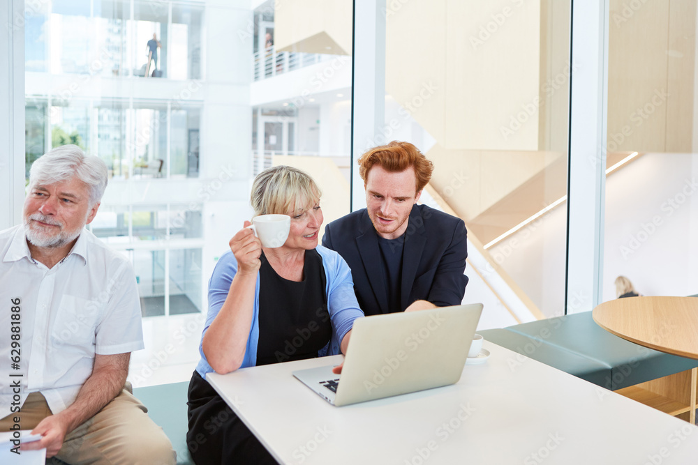 Mature businesswoman holding coffee cup while discussing over laptop with businessman by colleague in office lounge Mature businesswoman holding coffee cup while discussing over laptop with businessman by colleague in office lounge