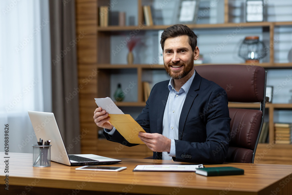 Portrait of happy businessman investor, mature adult man smiling and looking at camera, boss holding message envelope with good winning news inside office at workplace. Portrait of happy businessman investor, mature adult man smiling and looking at camera, boss holding message envelope with good winning news inside office at workplace.