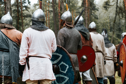 Back view of group of ancient warriors in chain mail, helmets with weapons standing in forest, costumed medieval festival outdoors