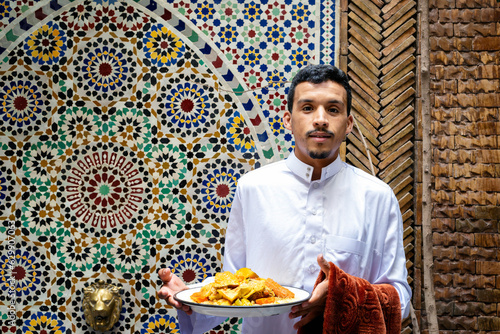 Portrait of a young muslim Berber man wearing a traditional white qamis and holding a vegetarian couscous plate. Marrakesh or Marrakech city. Morocco people.