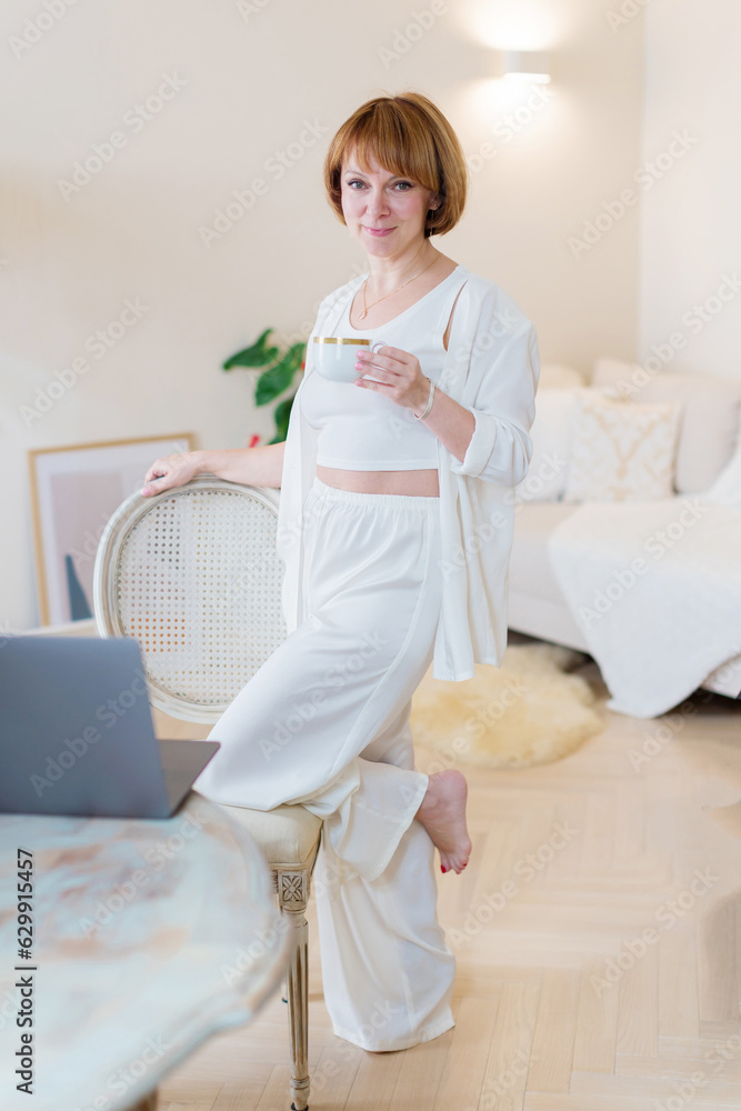 Happy smiling young woman taking a coffee break as she stands at her laptop computer working from a home office in stylish white livingroom. Happy smiling young woman taking a coffee break as she stands at her laptop computer working from a home office in stylish white livingroom.