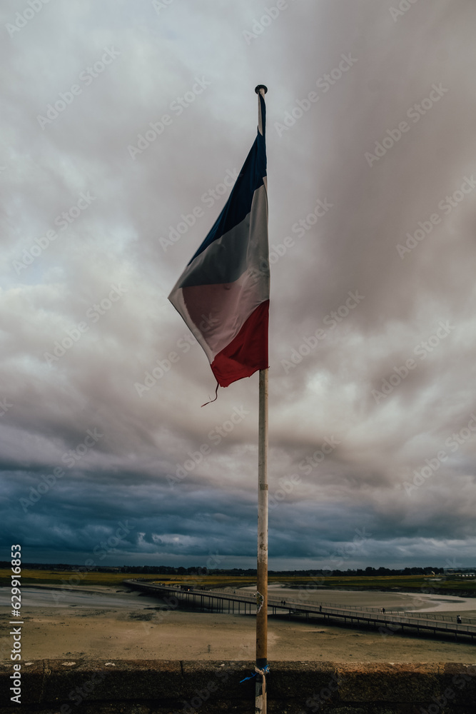 Foto vertical de la bandera francesa con un paisaje nublado al fondo desde Mont Saint Michel, Francia.