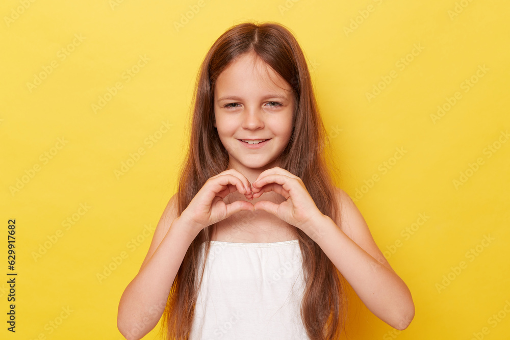 Smiling beautiful little girl with long hair standing isolated over yellow background expressing love showing heart shape with hands looking at camera with happy face. Smiling beautiful little girl with long hair standing isolated over yellow background expressing love showing heart shape with hands looking at camera with happy face.