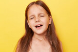 Closeup portrait of beautiful little girl with long hair standing isolated over yellow background standing with closed eyes pretending she crying