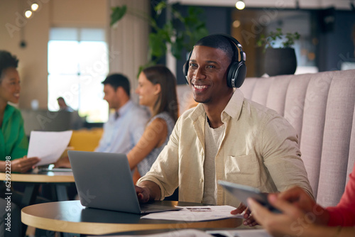 Businessman Wearing Wireless Headphones Working On Laptop In Informal Seating Area Of Modern Office