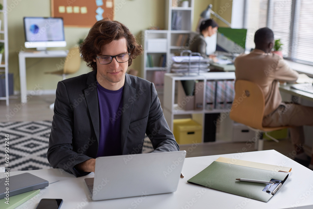 Portrait of young long haired businessman wearing glasses using laptop at office workplace and smiling, copy space