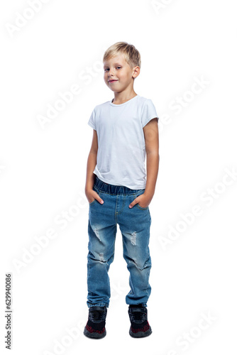Smiling boy stands with his hands in his pockets. A child in jeans and a white T-shirt. Positivity and confidence. Isolated on white background. Vertical.