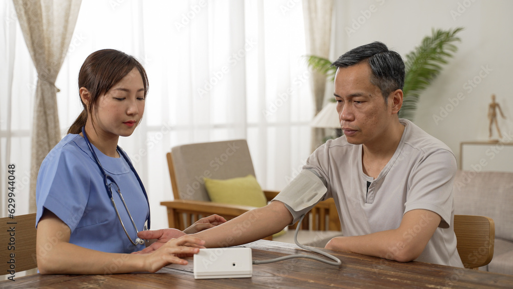 Asian female personal aide measuring blood pressure for the male elderly senior at home. she puts cuff on his upper arm and operates the device, waiting for result Asian female personal aide measuring blood pressure for the male elderly senior at home. she puts cuff on his upper arm and operates the device, waiting for result
