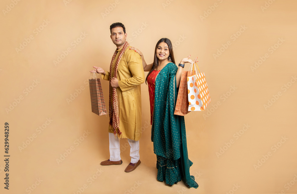 Happy young indian couple wearing traditional outfit holding shopping bags standing isolated on beige studio background. Diwali celebration and festive sale concept. Full length. Copy space.