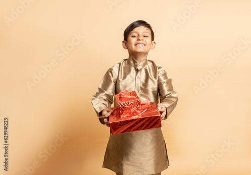 Happy young indian kid boy wearing traditional kurta outfit holding red gift boxes celebrating of diwali festival isolated on beige background. Child holding present.