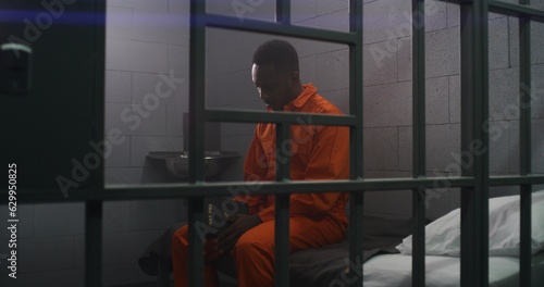African American prisoner in orange uniform sits on the bed behind bars, reads Bible in prison cell. Criminal serves imprisonment term for crime in jail. Detention center or correctional facility.