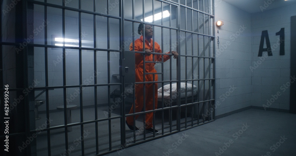 African American prisoner in orange uniform leans on jail cell bars and looks around. Guilty gangster serves imprisonment term in correctional facility. Gloomy inmate in detention center.