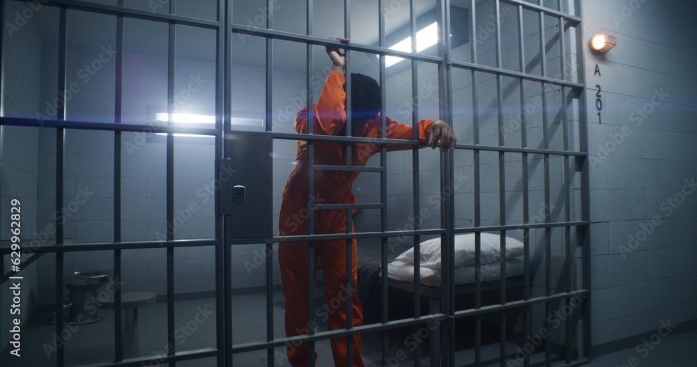 African American prisoner in orange uniform leans on jail cell bars and ...