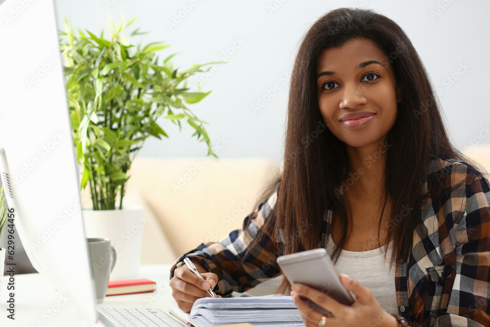 Smiling young african american woman writing in notebook studying on mobile phone online. Happy ethnic woman taking handwritten notes on paper with smartphone app in hand
