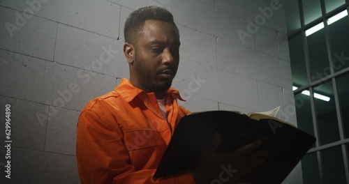 African American prisoner in orange uniform sits in prison cell, reads Bible. Male criminal serves imprisonment term for crime in jail. Offender in detention center or correctional facility. Portrait.