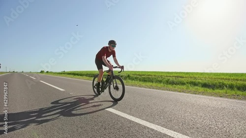 Fully-equipped focused male changing standing up position on bike into seated one after acceleration. Experienced rider in sports apparel performing cycling workout on cleared roadway in summer.