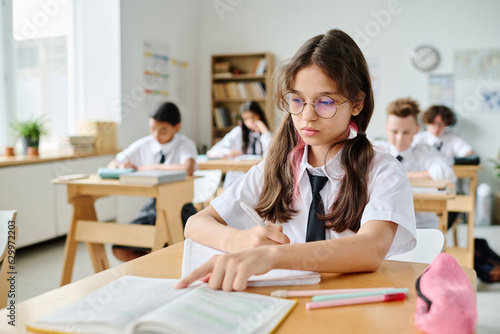 Schoolgirl sitting at her desk and reading textbook during lesson in school