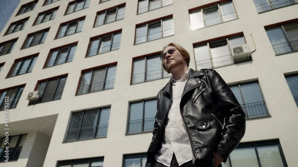 A man in sunglasses stands against the background of a building and looks into a wristwatch. Modern wristwatches. Sunglasses and black leather jacket. A man in sunglasses stands against the background of a building and looks into a wristwatch. Modern wristwatches. Sunglasses and black leather jacket.