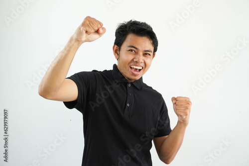 smiling or happy young asian man clenched his fists cheering carefree and excited. Victory concept, wearing black polo t shirt isolated on white background