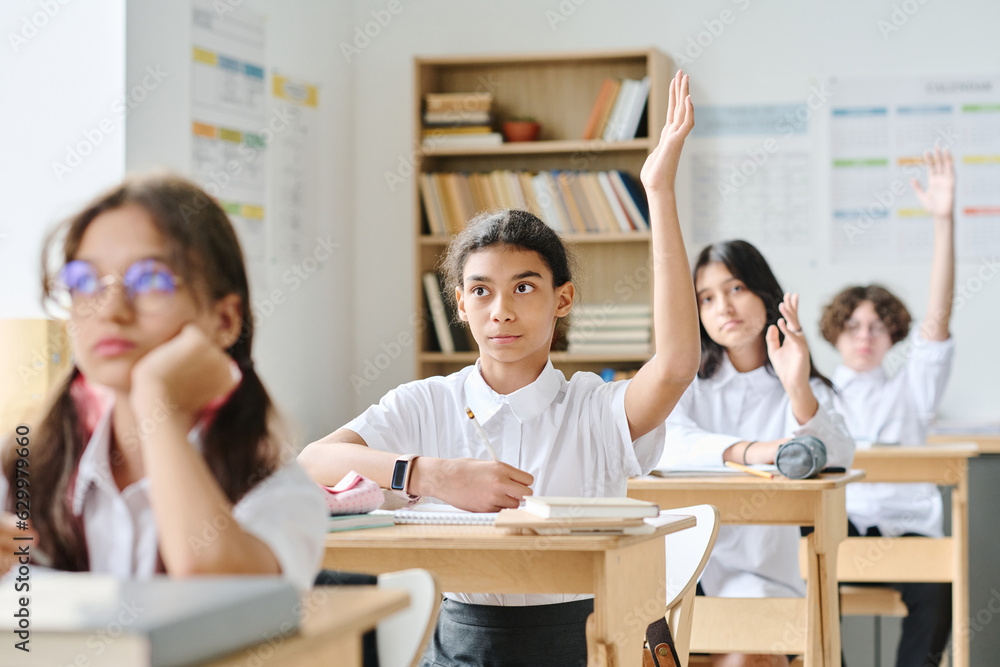 Schoolgirl sitting at her desk and raising her arm, she answering at lesson at school Schoolgirl sitting at her desk and raising her arm, she answering at lesson at school
