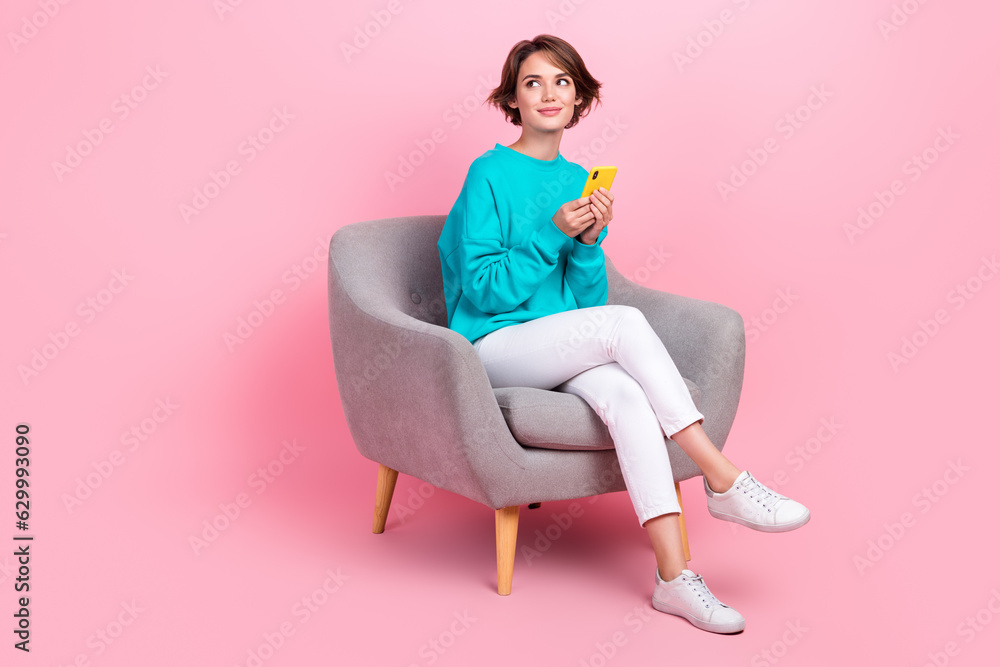 Full body photo of attractive young woman armchair hold device look empty space wear trendy blue clothes isolated on pink color background Full body photo of attractive young woman armchair hold device look empty space wear trendy blue clothes isolated on pink color background