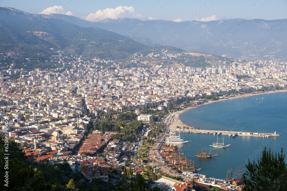 Enjoy the stunning view of Alanya from the observation deck, with lush trees, blue sea, and the iconic Lighthouse and Port of Alanya. The rocky peninsula adds a dramatic touch to the panoramic scene.
