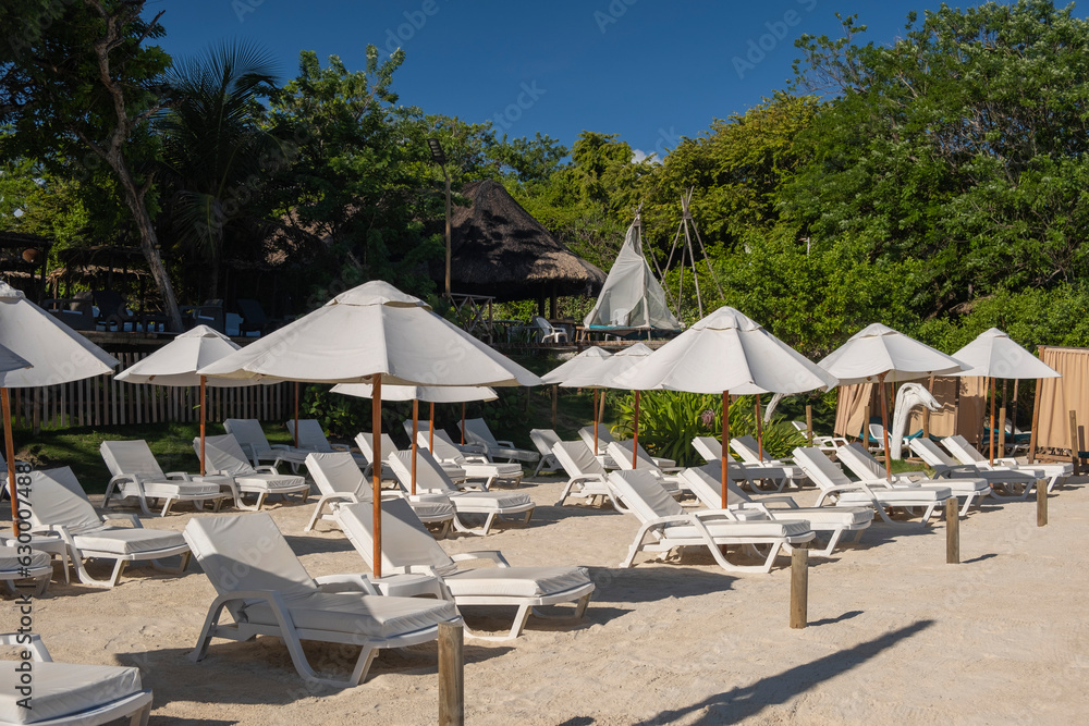 Beach scene - rows of lounge chairs and sun umbrellas on the white sand beach with no people, tourists or travelers around surrounded by lush green trees and palms. Clear blue sky, sunny day.