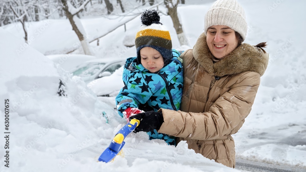 Smiling mother and her baby son cleaning snow from their car with a brush after a blizzard on a parking lot. The concept of daily routine and taking care of their vehicle