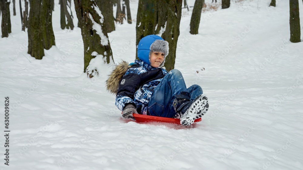 Happy smiling boy riding down the snowy hill on plastic sleds. Concept of winter holiday, children having fun and playing outdoors in snow, Christmas vacation. Happy smiling boy riding down the snowy hill on plastic sleds. Concept of winter holiday, children having fun and playing outdoors in snow, Christmas vacation.