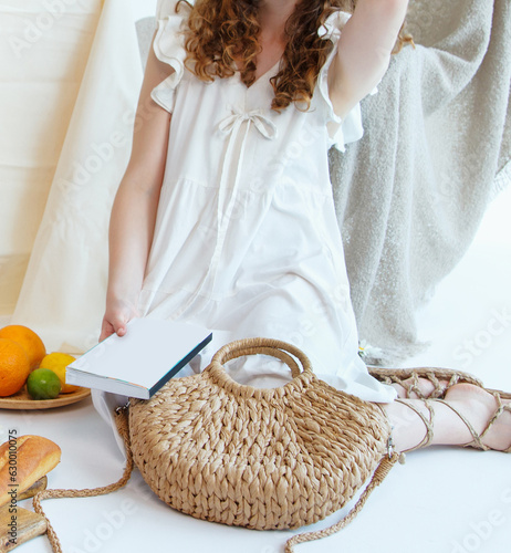 Summer fashion eco style. A girl in a white dress sits on the floor with a bag on her lap  wrapped in sandals made of natural materials and holding a book in her hand. Nearby lie fruit on plates. 