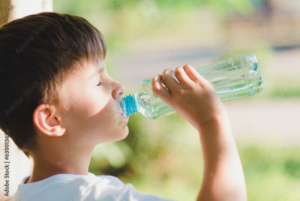 Cute boy sitting on the grass drinks water from a bottle in the summer at sunset. Child quenches thirst on a hot day Cute boy sitting on the grass drinks water from a bottle in the summer at sunset. Child quenches thirst on a hot day