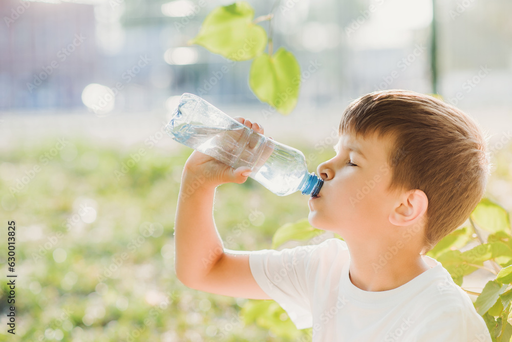 A beautiful child sitting on the grass drinks water from a bottle in the summer at sunset. Boy quenches his thirst on a hot day A beautiful child sitting on the grass drinks water from a bottle in the summer at sunset. Boy quenches his thirst on a hot day
