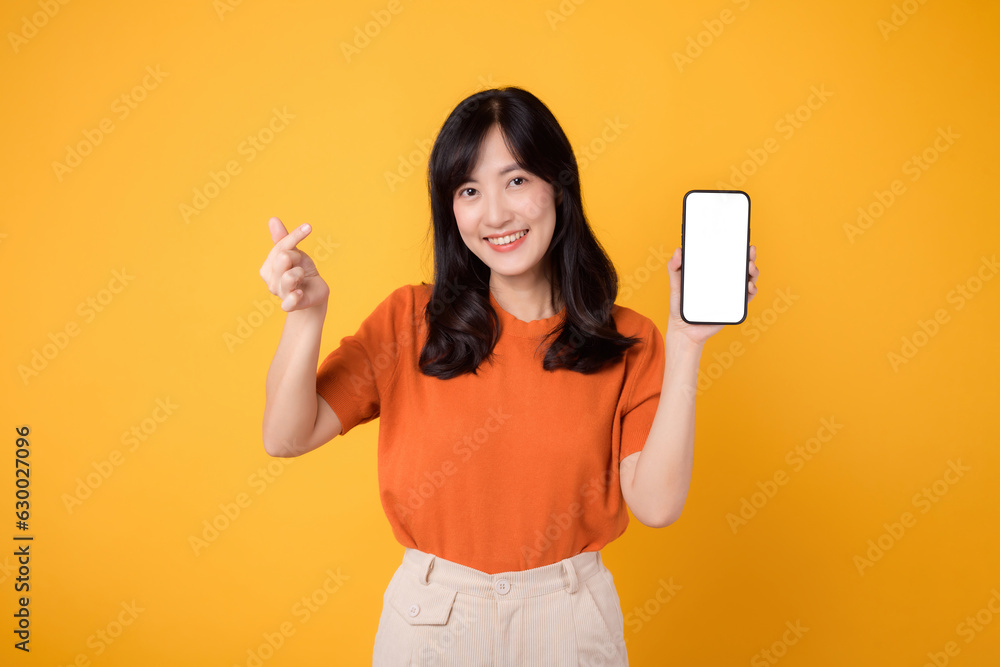 Cheerful young Asian woman in her 30s, wearing an orange shirt, showing mini heart hand gesture and smartphone blank display screen on yellow background. new application technology concept. Cheerful young Asian woman in her 30s, wearing an orange shirt, showing mini heart hand gesture and smartphone blank display screen on yellow background. new application technology concept.