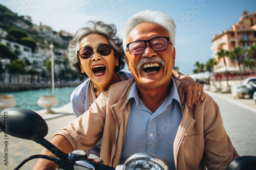 Wearing smiles mirror their carefree spirit, the Asian seniors ride scooter, possibly exploring picturesque coastal town, feeling the joy of the wind in their hair.