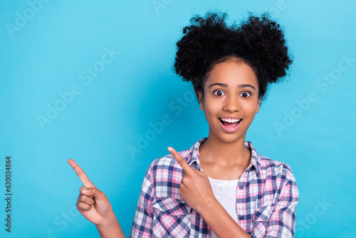 Photo of impressed astonished girl with buns hairstyle dressed checkered shirt directing empty space isolated on blue color background