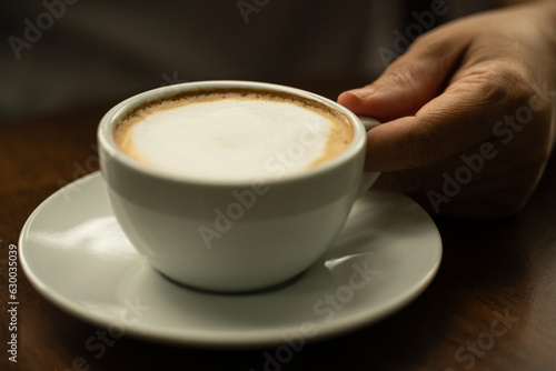 Fototapeta Naklejka Na Ścianę i Meble -  Closeup hand holding white cup of hot coffee latte on wood background in restaurant.Best of menu in the coffee shop.Soft focus.