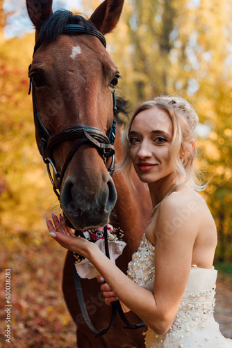 Beautiful bride in white dress stands near a horse in the autumn forest.