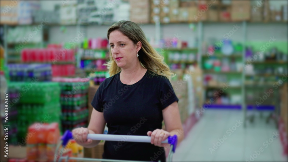 Fototapeta premium Middle-Aged Caucasian Woman Shopping in Supermarket Aisle with Cart, Examining Products