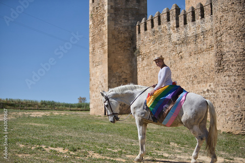 Non-binary person, young and South American, very makeup, mounted on a white horse, smiling and happy, with a gay pride flag on the rump, next to an old medieval castle. Concept queen, lgbtq+, pride. © Manuel