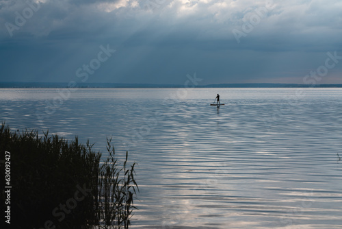 young man standing on sup and paddling on water of calm lake, tranquil scenery
